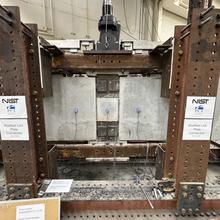 A wide view of a concrete beam assembly inside a large metal frame. A hydraulic arm is part of the metal frame, and it’s posed to press down at a section in the middle where the concrete pieces connect.
