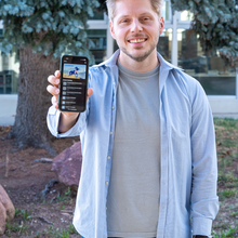 Bill Fisher smiles as he poses holding out a smartphone that shows a mobile driver's license. 