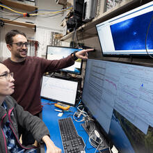 A woman with glasses and a long ponytail is seated at a computer with four display screens. A man with glasses stands beside her and points at the uppermost display screen. In the middle of that screen is a small blue glowing dot. The lower screens display data from the experiment.