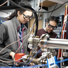 Two researchers wearing safety glasses are seen standing in the lab surrounded by wiring and electronics.