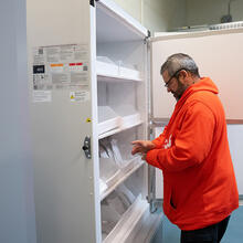A man in a bright orange sweatshirt stands at the open door of a large lab freezer.