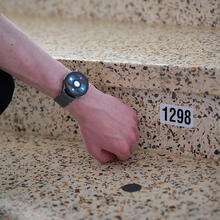 A person wearing a specialized smartwatch gestures to a black dot sticker with a nearby numbered sticker on a set of stairs.