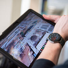 A person wearing a specialized smartwatch holds a tablet showing a photo of the NIST campus with colored dots marking certain locations. 