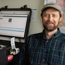 Gary Howarth poses for a photo in his office, wearing a NIST baseball cap, with a ruggedized laptop on the counter next to him and a map on the wall behind him.. 