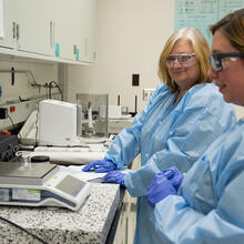 Two women in blue paper lab coats and safety glasses stand at a lab counter, looking at a small glass container on a scale. 