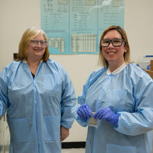 Two women in blue paper lab coats and safety glasses stand smiling in the lab. 