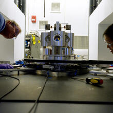 Wide shot shows two researchers on either side of a scientific device made of metal cylinders, peering closely at it. 
