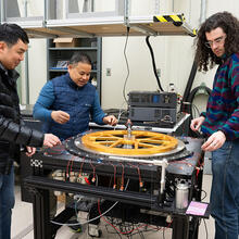Three warmly dressed researchers stand around a scientific device with a large circular piece lying flat on top. 