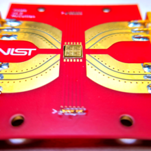 A bright-red rectangle with a NIST logo holds a gold-colored chip in the center with metal paths leading to connectors at the edge of the rectangle. 