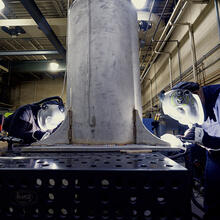 Two people wearing welding masks and other safety equipment bend over a table to work on a large metal cylinder.