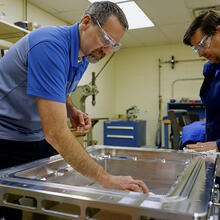 Two researchers wearing safety glasses lean over a rectangular metal case lying flat on a table.