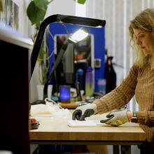 A woman wearing special gloves stands at a high table, sorting small pieces of paper. 