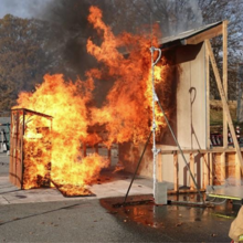 A very small wooden shed filled with 6 wood cribs, separated by 3.2 m (10 ft) from the target, and burning under a high wind condition creates a large plume of flame that contacts the target wall.