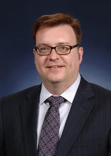 Headshot of Mike Fasolka, a man with short brown hair, wearing glasses, a dark blue suit jacket, white dress shirt, and purple decorated tie