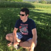 Photo of a man wearing short sleeves, shorts, and sunglasses sitting in the grass