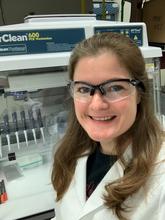 Megan Cleveland with white lab coat and safety glasses in front of a lab bench with pipettes. 