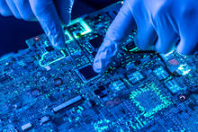 Closeup photo shows person wearing blue gloves using tweezers to work on a semiconductor circuit board.