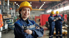 A woman in a construction hat stands smiling with her arms crossed in a factory work environment. 