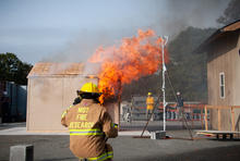 Photo shows a NIST firefighter on standby watching a shed burn next to a target structure representing a wall of a residence during an experiment.