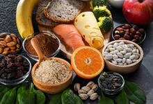 Photograph of a collection of different foods including bread, fish, cheese, fruits, nuts, and grains