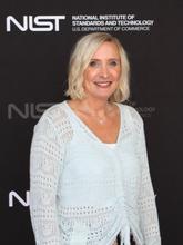 A woman with short blonde hair and a light blue blouse, standing in front of a NIST photo backdrop