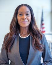 Headshot of a woman in a gray suit with pearls. Blurry American flag in the background