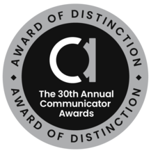 Circle badge. Silver exterior ring with writing at top and bottom: Award of Distinction. Black interior. Lowercase letter A. Words: The 30th Annual Communicator Awards
