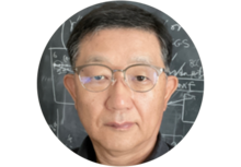 person with short black hair and wearing glasses, standing in front of a chalk board covered in calculations