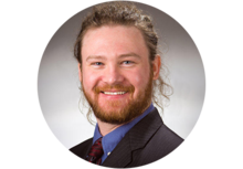 person with a curly brown ponytail and a beard, wearing a suit 