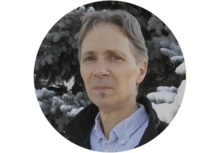 person with short grey hair wearing a dress shirt, standing in front of snow-covered evergreens