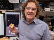 NIST's Andrew Wilson holds an ion trap in his lab in Boulder, Colorado. Behind him are screens depicting dots.
