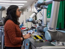  A person with long black hair holds a tablet and looks up at a robot, while working in a lab at NIST. 