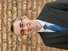 Samuel Hales, a young man with short blonde hair and glasses, wearing a suit and tie, standing in front of a brick wall.
