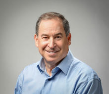 A man with short gray hair, wearing a blue collared shirt, smiling.