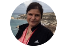 Headshot of Suzanne Lightman, a woman with dark hair, pulled back, wearing a pink shirt and black jacket, in front of rocky beach scene.