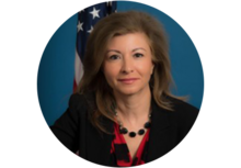 Professional headshot of Azita Valinia, a woman with long brown hair, wearing a necklace, black blazer, and red top, smiling in front of a U.S. flag.