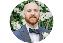 Professional headshot of Justin Fessler, a bald man with a mustache and beard, wearing a bow tie and suit, smiling outside in front of flowers.