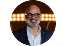 Professional headshot of Quintin D. Jones, a bald man with a mustache, beard, and glasses, wearing a collared button up shirt and suit jacket, with studio lights in the background.
