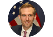 Professional headshot of Taylor Jordan, a man with short blonde hair, smiling, wearing a suit and tie, in front of an American flag background.