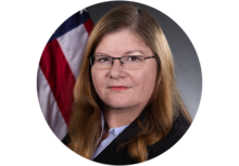 Professional headshot with American flag in the background featuring Melissa Vice, a woman with long dirty blonde hair and glasses, wearing a blazer.