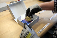 A gloved hand holds a commemorative coin featuring Theodore Roosevelt over an open white box. 