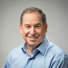 A man with short gray hair, wearing a blue collared shirt, smiling. 