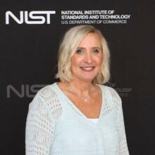 A woman with short blonde hair and a light blue blouse, standing in front of a NIST photo backdrop.