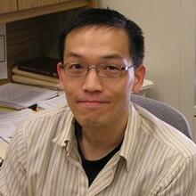 A man with short black hair, wearing a glasses, sitting at a desk, and smiling at the camera.