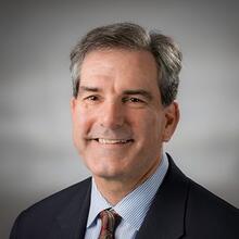 Professional headshot of Steven Choquette, a man with short black-gray hair, in a blue striped collared shirt, dark suit jacket, and a decorated tie.