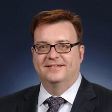 Headshot of Mike Fasolka, a man with short brown hair, wearing glasses, a dark blue suit jacket, white dress shirt, and purple decorated tie
