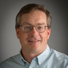 Professional headshot of John Perkins, a man with short brown-gray hair, wearing glasses, a blue collared shirt, and smiling.