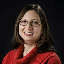 Rebecca Zangmeister, a woman with shoulder length dark brown hair and glasses, smiling for a professional headshot.