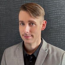 Headshot of Kyle Anderson, a person with short blonde hair, wearing a black collared shirt and a gray suit jacket, smiling