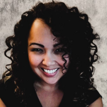 Nathali Clark, a person with long curly black hair, smiling for a headshot against a gray stone background. 